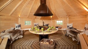 Interior de una cabaña de madera con una mesa central dispuesta para comer, rodeada de alfombras de piel y una cesta grande de mimbre.
