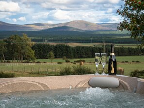 A hot tub with a bottle and two glasses, surrounded by a scenic landscape with mountains and fields. - The Eagle's Nest - large luxury lodge with hot tub, sauna and bbq hut (Boat of Garten)