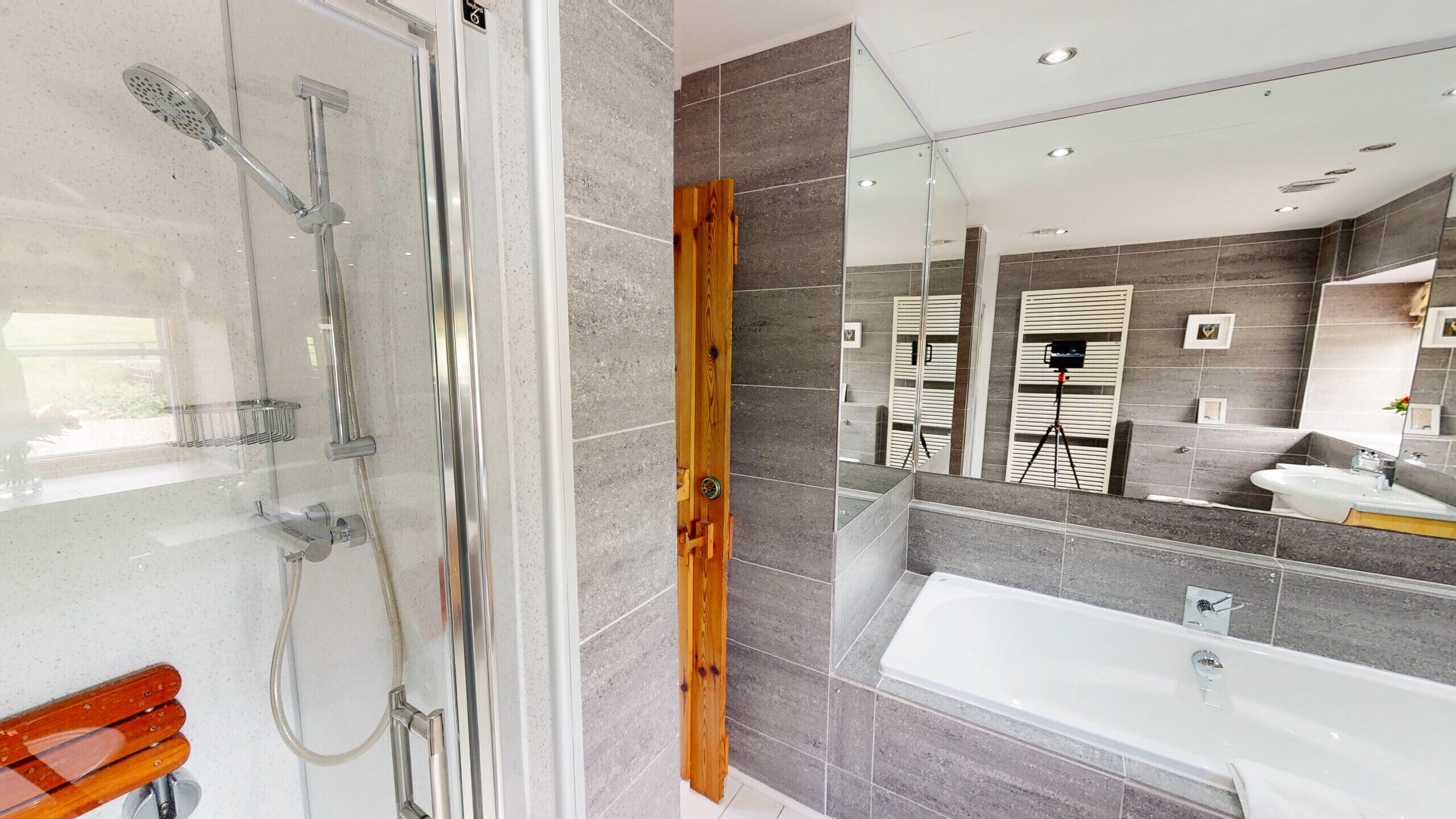 A modern bathroom with a glass-enclosed shower, a wooden door, a large mirror, and a white bathtub.