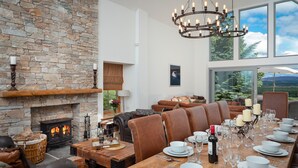 Un comedor con una larga mesa de madera dispuesta para una comida, una chimenea de piedra, asientos de cuero y un amplio ventanal con vistas panorámicas.