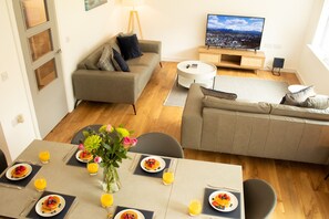 Un salon moderne avec un canapé, une table à manger dressée pour le petit-déjeuner, un téléviseur et une vue sur le paysage urbain.