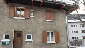 Un edificio de dos pisos con fachada de hormigón, varias ventanas con contraventanas de madera y una puerta con un pequeño ventanal encima.