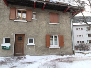 Edificio a due piani con facciata in cemento, diverse finestre con persiane in legno e una porta con un piccolo oblò sopra.