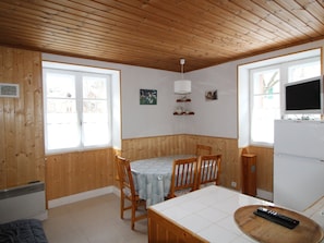 A cozy room with wooden walls and a tiled floor, featuring a dining table with chairs, a small TV, and a refrigerator.