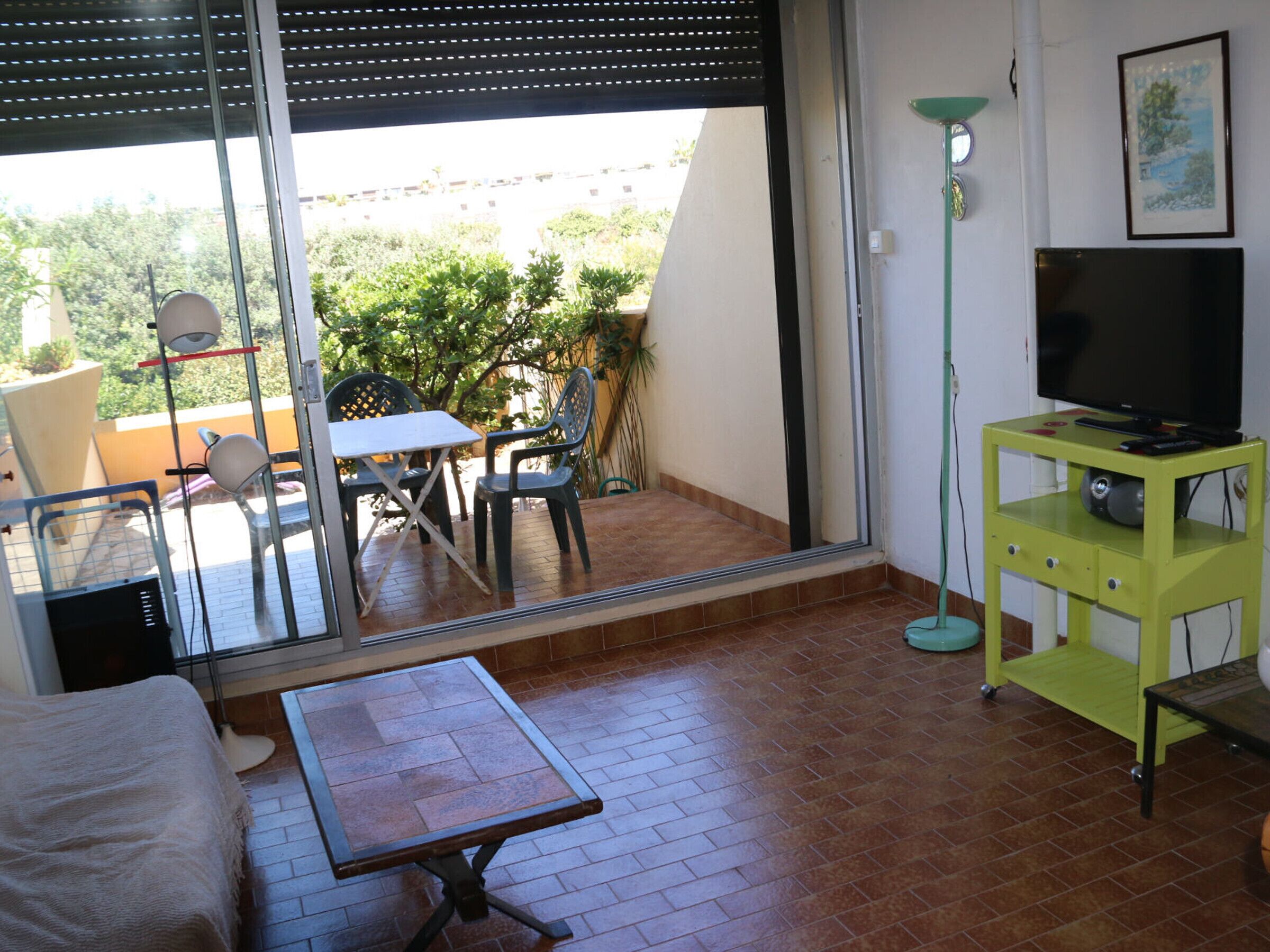 Un salon comprenant un canapé, une petite table, un téléviseur posé sur un meuble, et un balcon avec une table et des chaises.
