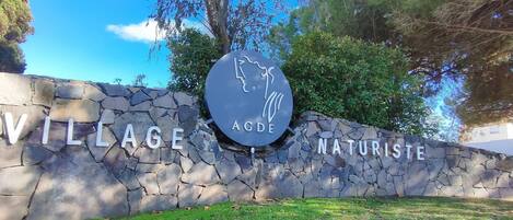 A stone wall with the words "VILLAGE AODE NATURISTE" and a circular logo with a handprint.