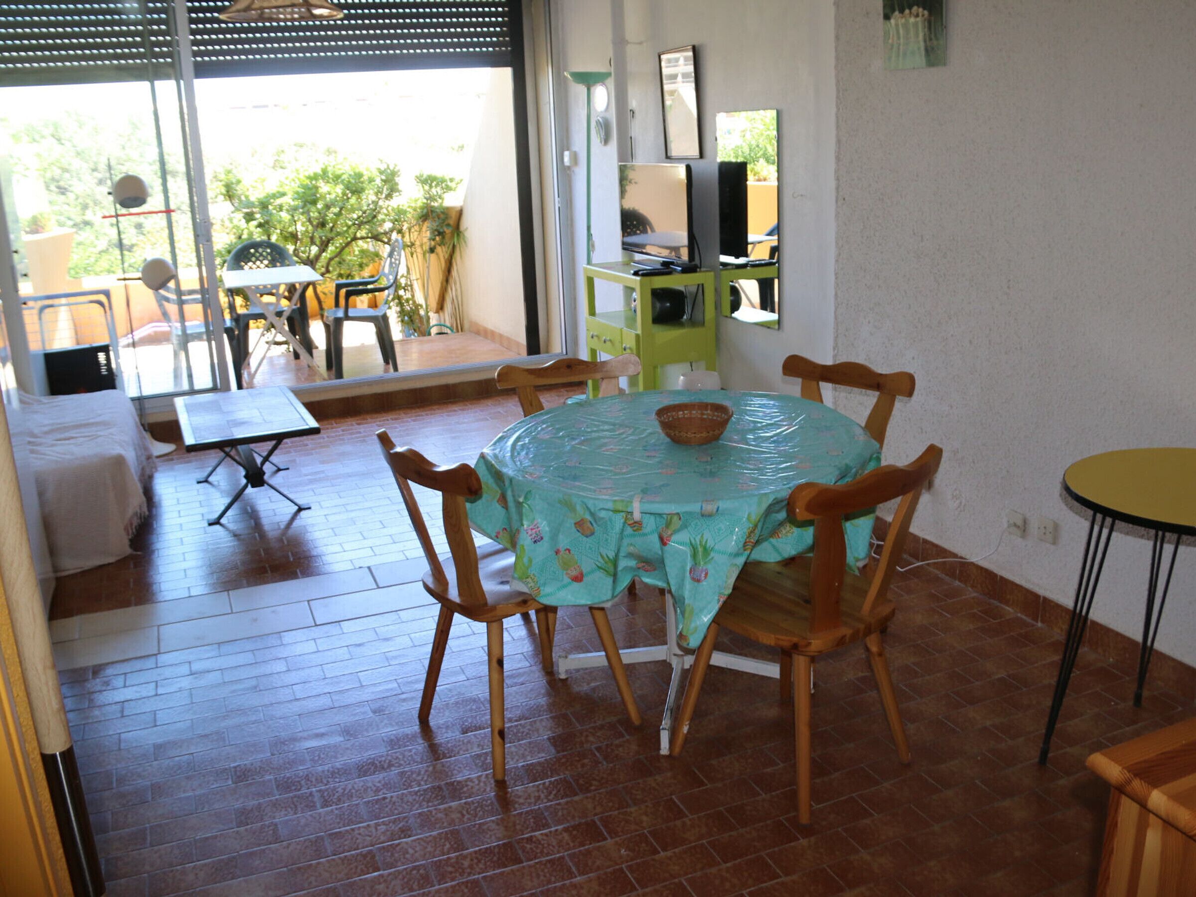 A dining area with a round table covered by a tablecloth, surrounded by wooden chairs. There is a small table with a chair outside, and a bench with a cushion is visible in the background.