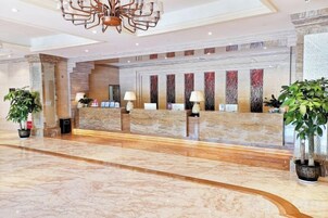 A hotel reception area with marble flooring, a reception desk, potted plants, and a chandelier.