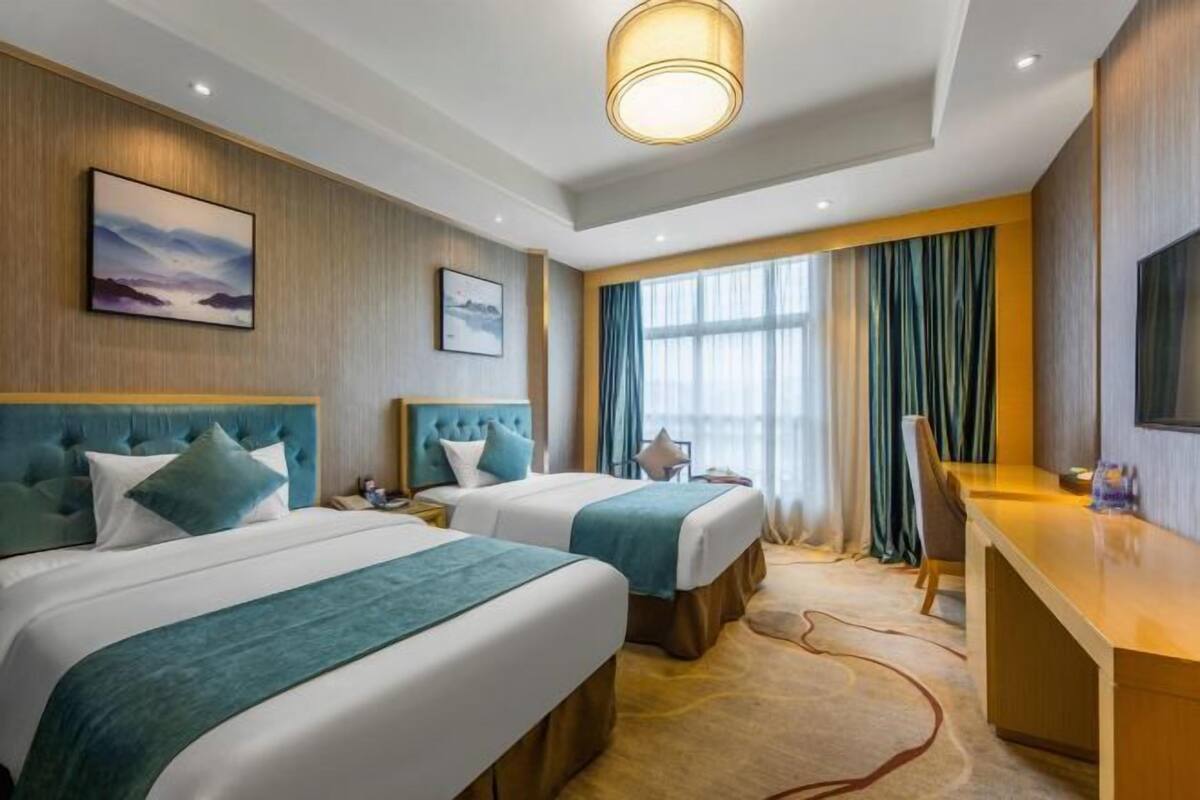A hotel room with two beds, a desk, a large window with curtains, and artwork on the walls.