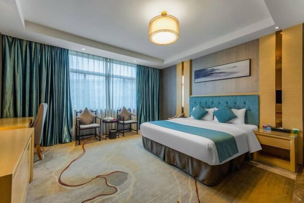 A hotel room with a large bed, a desk, two chairs, a desk lamp, and a painting on the wall.