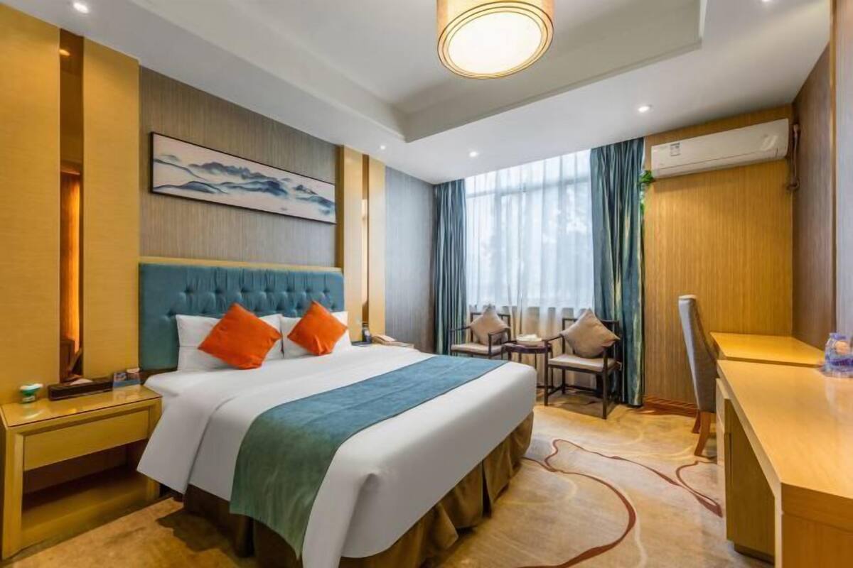A hotel room with a large bed, a desk, and a chair. There is a painting on the wall and an air conditioning unit on the wall.
