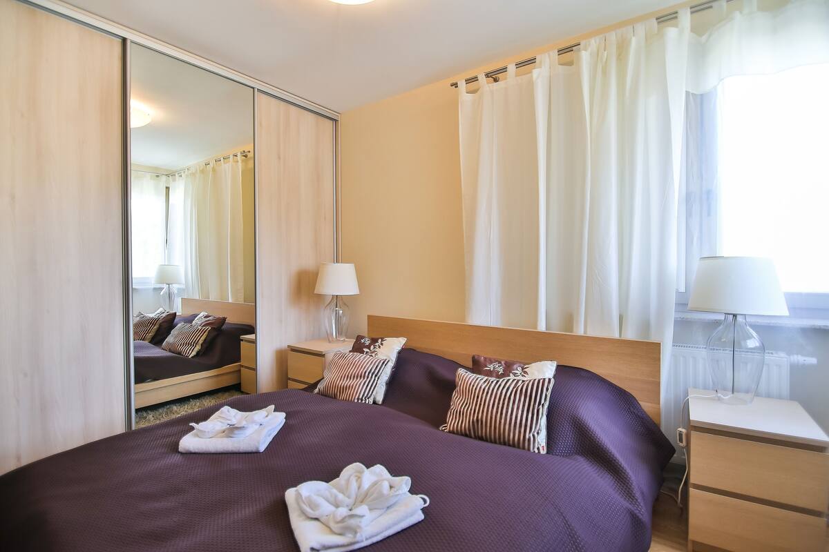 Ein Hotelzimmer mit einem großen Bett, einem Nachttisch, einem Spiegel und einem Fenster mit Vorhängen.