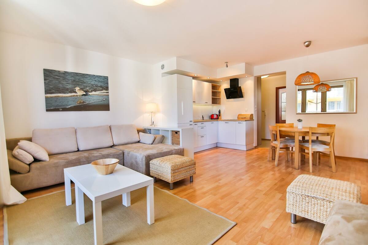 Ein modernes Wohnzimmer mit Sofa, Couchtisch und Hocker, eine Küche mit weißen Schränken und ein Essbereich mit Holzkreisen.
