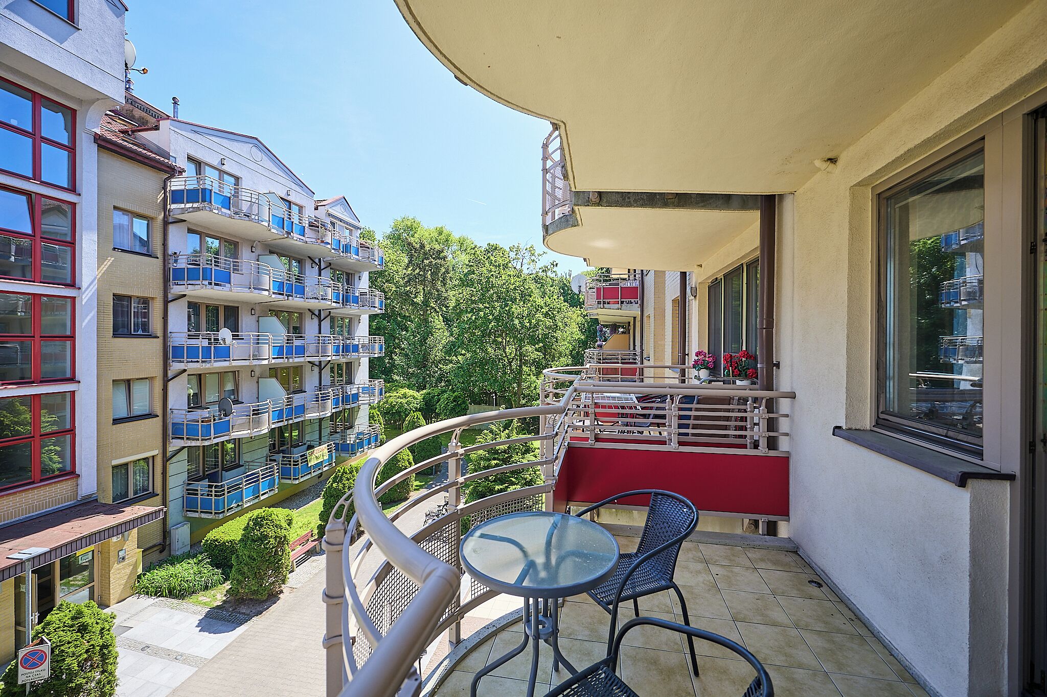 A balcony with a table and chairs, a red railing, and a view of neighboring buildings and trees.