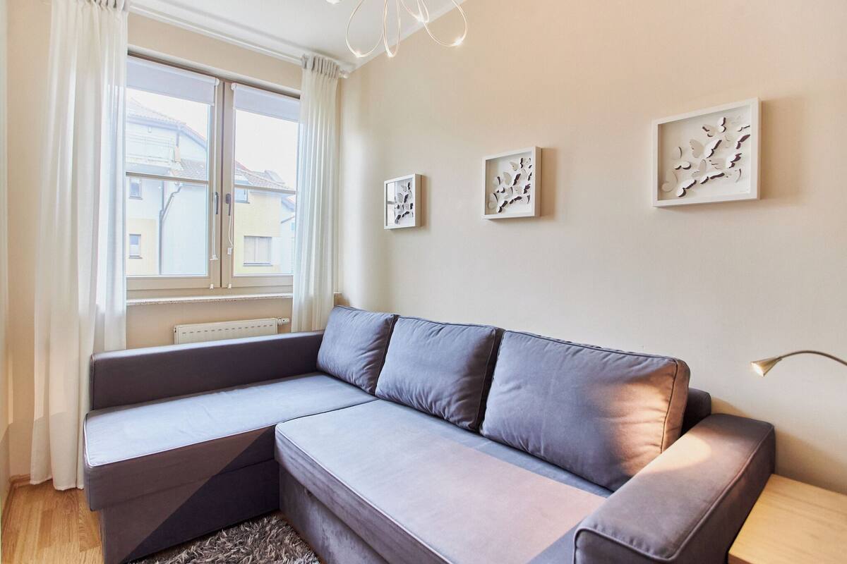 Ein modernes Wohnzimmer mit einem grauen Ecksofa, einem großen Fenster mit weißen Vorhängen und drei an der Wand befestigten abstrakten Kunstwerken.