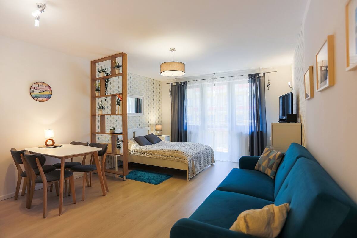 Ein modernes Schlafzimmer mit Bett, einem blauen Sofa, einem Esstisch mit Stühlen, einem kleinen Schreibtisch und einer Wanduhr.