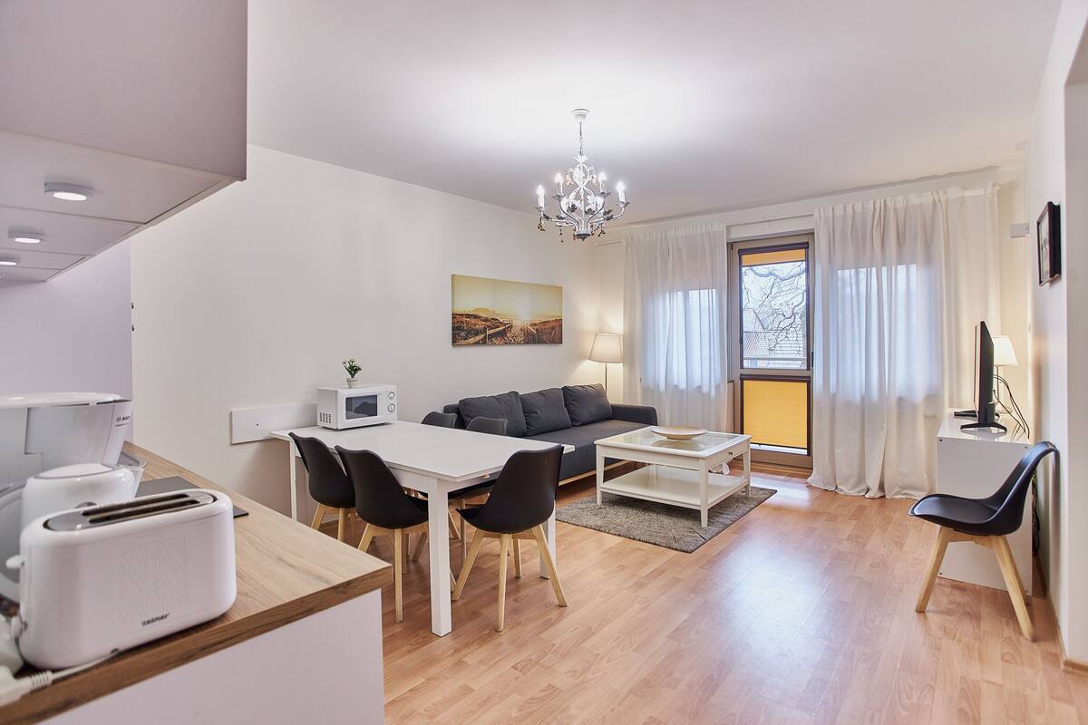 Ein modernes Wohnzimmer mit Essbereich, einer Couch, einer Chandelierleuchtre und Blick durch ein Fenster.
