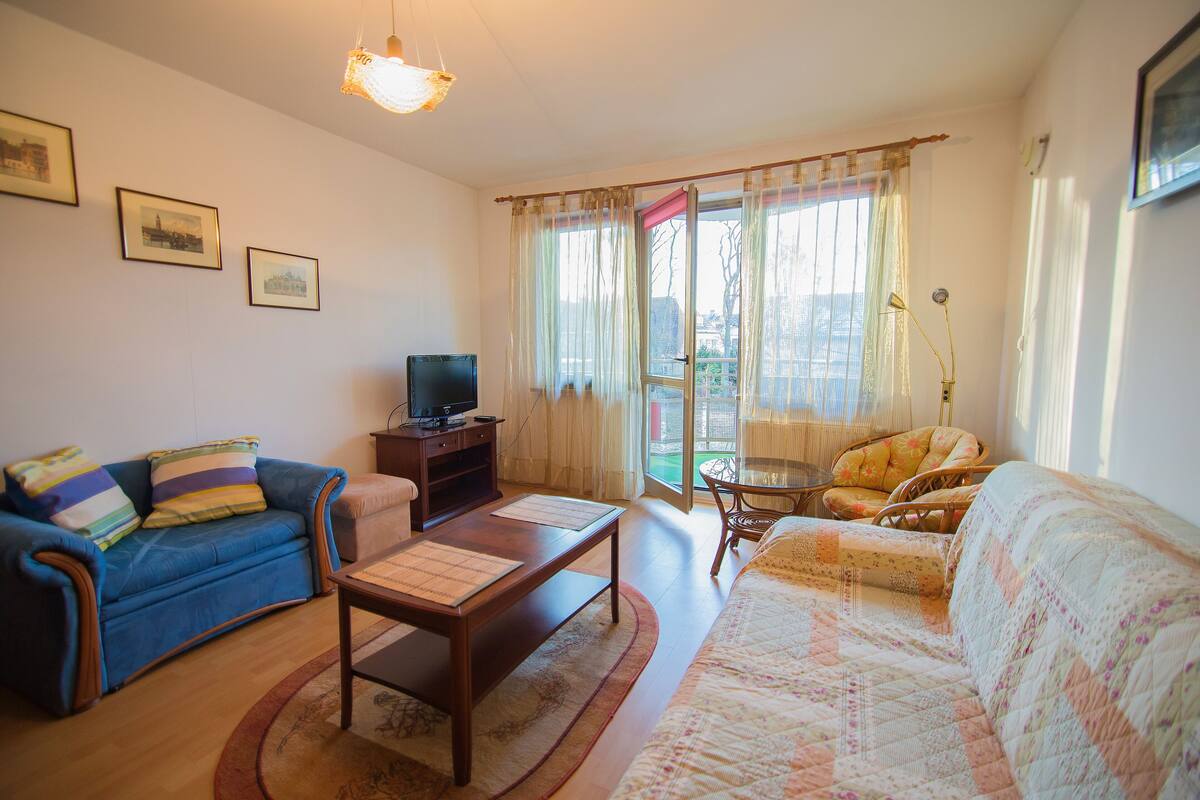 Ein Wohnzimmer mit blauem Sofa, Couchtisch aus Holz, einem Sofa mit Blumenmuster, einem Fernseher und einem Fenster mit Vorhängen.