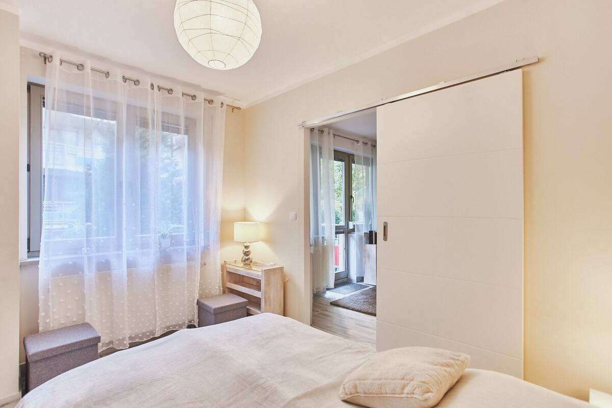 Ein Schlafzimmer mit einem Bett, einem Nachttisch mit Lampe, einem Fenster mit durchsichtigen Vorhängen und einer Schiebetür, die auf einen Balkon führt.