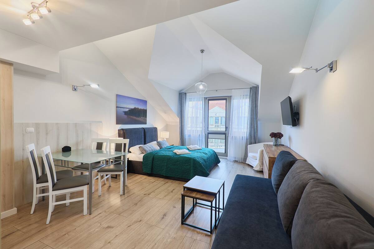 Ein modernes, gut beleuchtetes Zimmer mit Essbereich, einem Bett mit grüner Bettwäsche und einem großen Fenster.