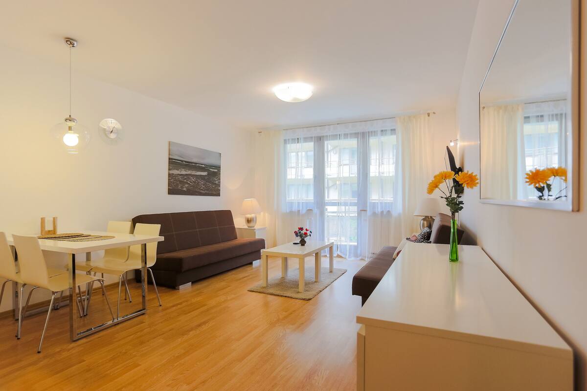 Ein modernes Wohnzimmer mit Sofa, Esstisch und Couchtisch, mit einem großen Fenster mit Vorhängen.