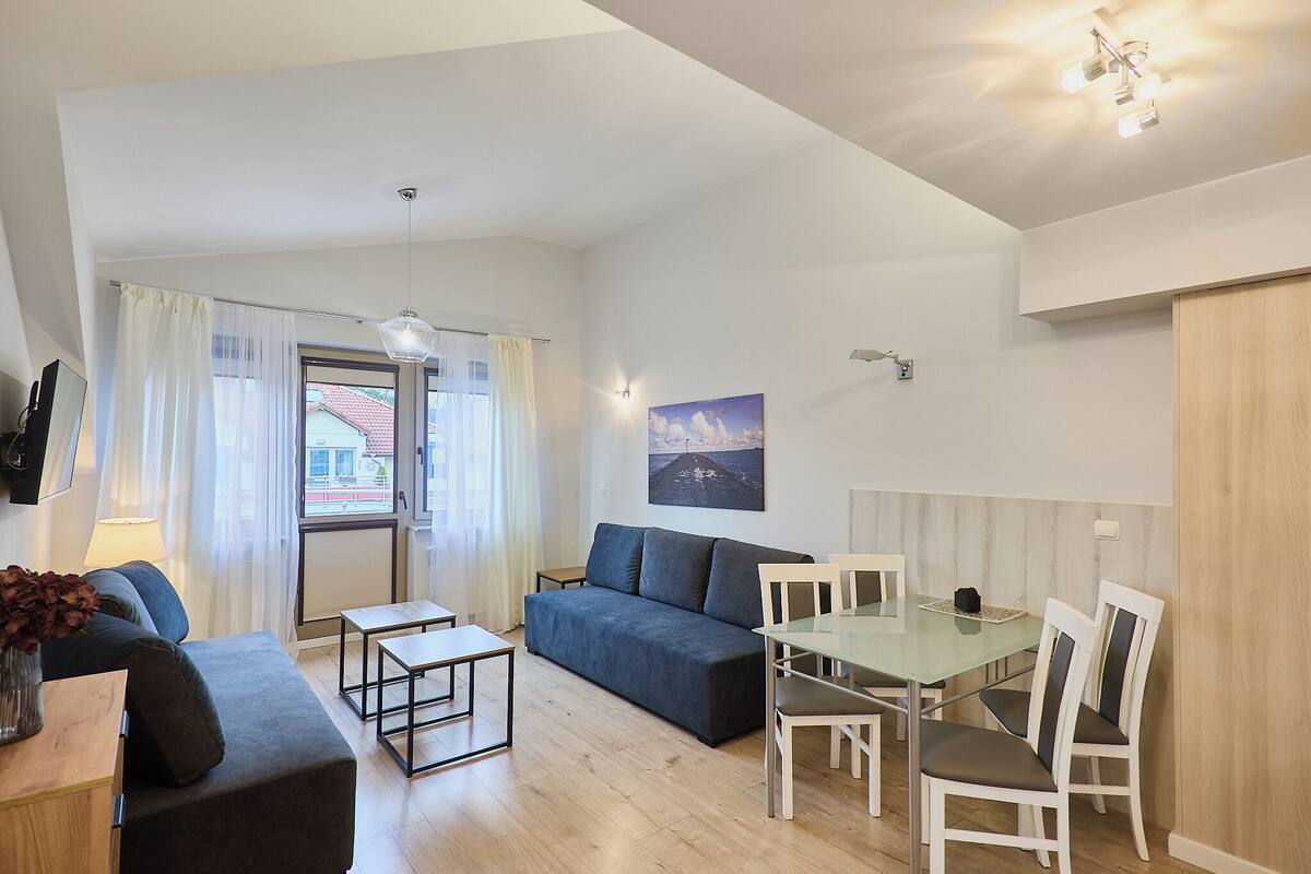 Ein modernes Wohnzimmer mit einer Couch, einem Glastisch und Stühlen. Es gibt ein Fenster mit Vorhängen, ein Bild an der Wand und eine Lampe.