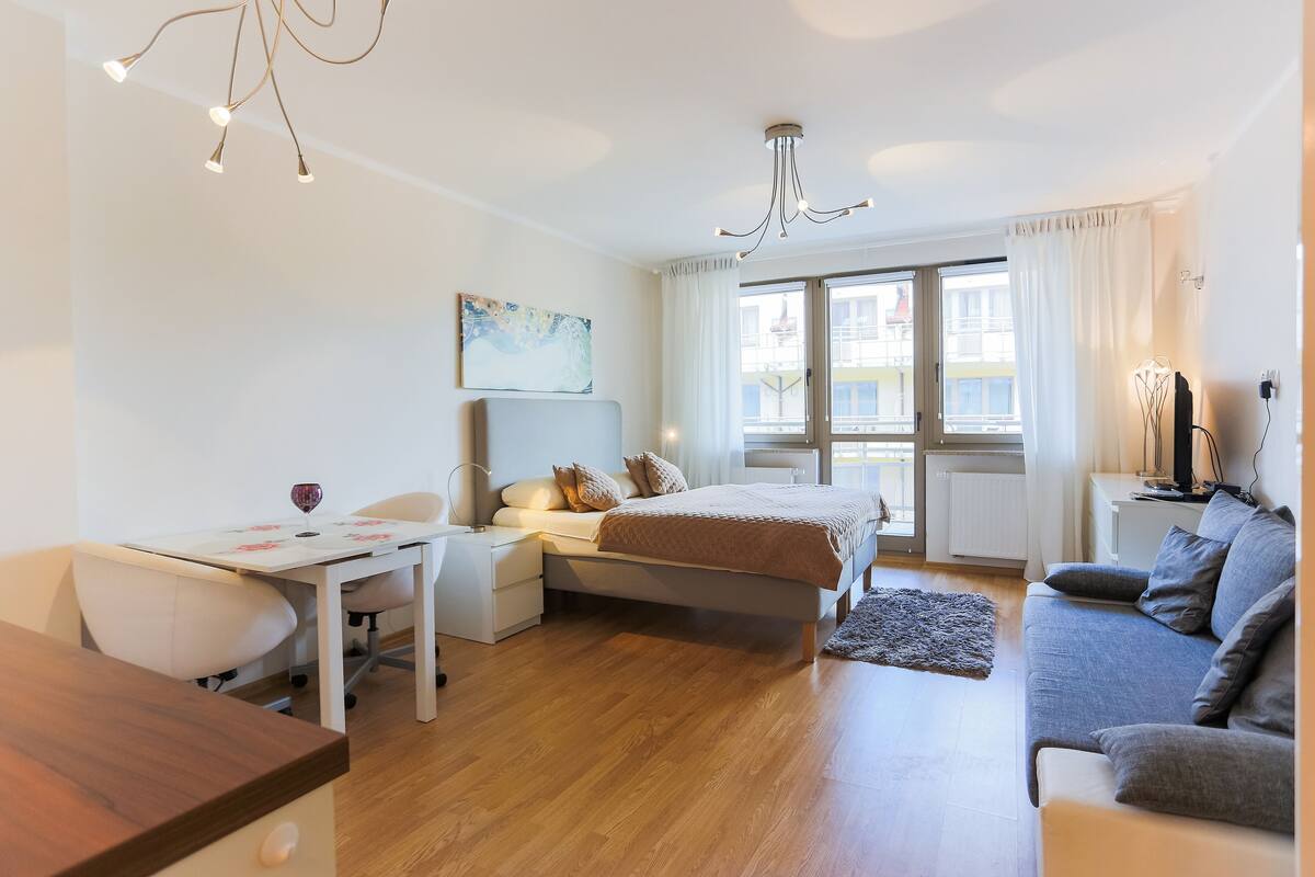 Ein modernes Schlafzimmer mit Bett, Nachttisch und Schreibtisch mit Stuhl. Ein Fenster mit Vorhängen bietet Blick auf Gebäude.