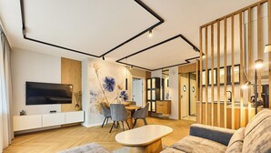 A modern living room with a flat-screen TV, a dining area with chairs, a wooden dining table, and a sofa.