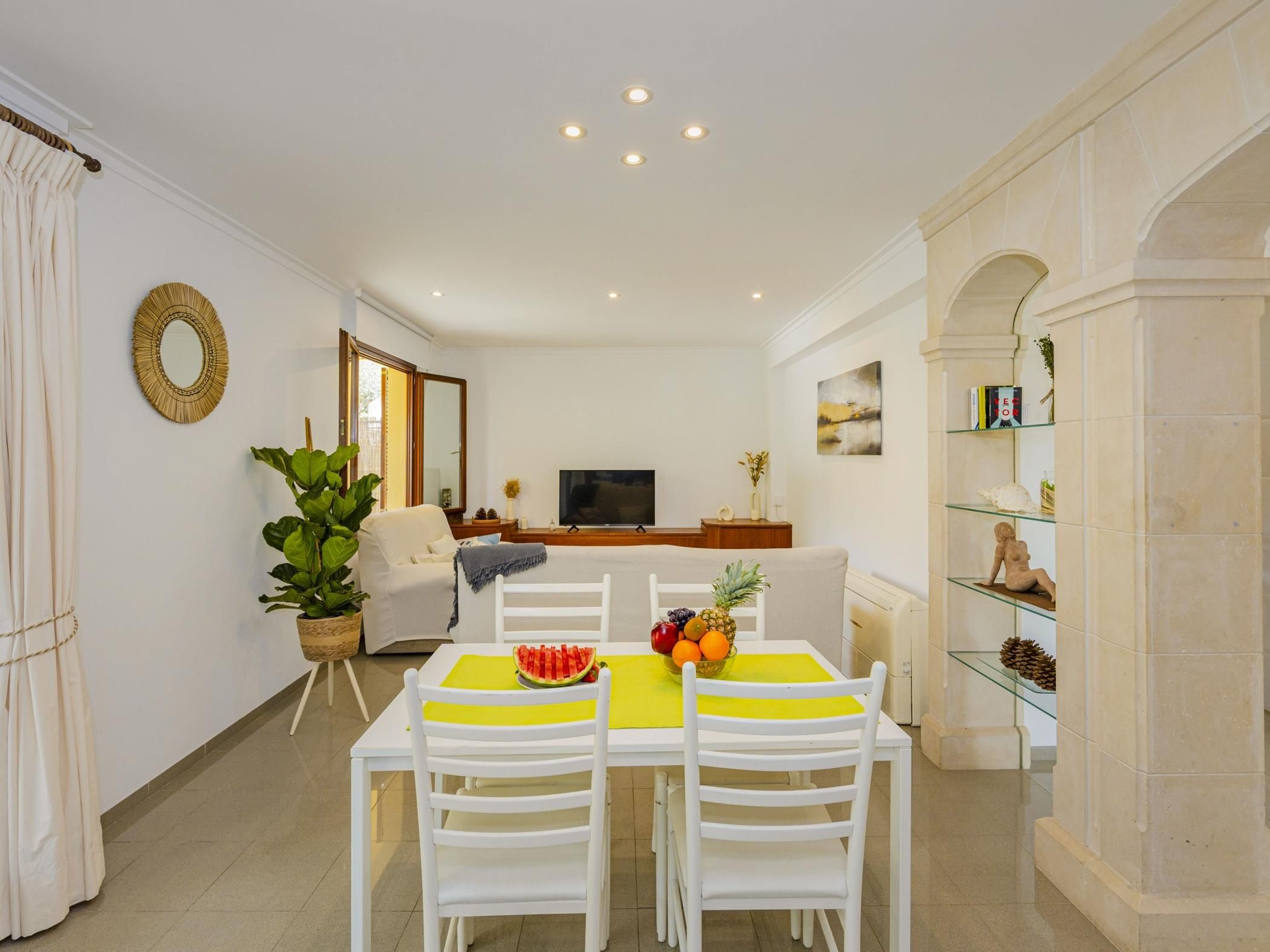 Un coin repas avec une table dressée pour deux personnes, un téléviseur en arrière-plan et une plante sur la gauche.