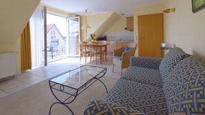 A modern living room with a glass coffee table, a patterned sofa, and a dining area with wooden chairs and a kitchen in the background.