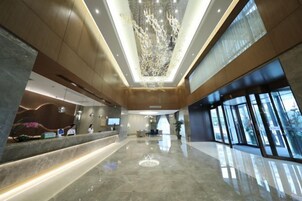 A spacious lobby with a reception desk, a high ceiling with a decorative light fixture, and large windows allowing natural light.