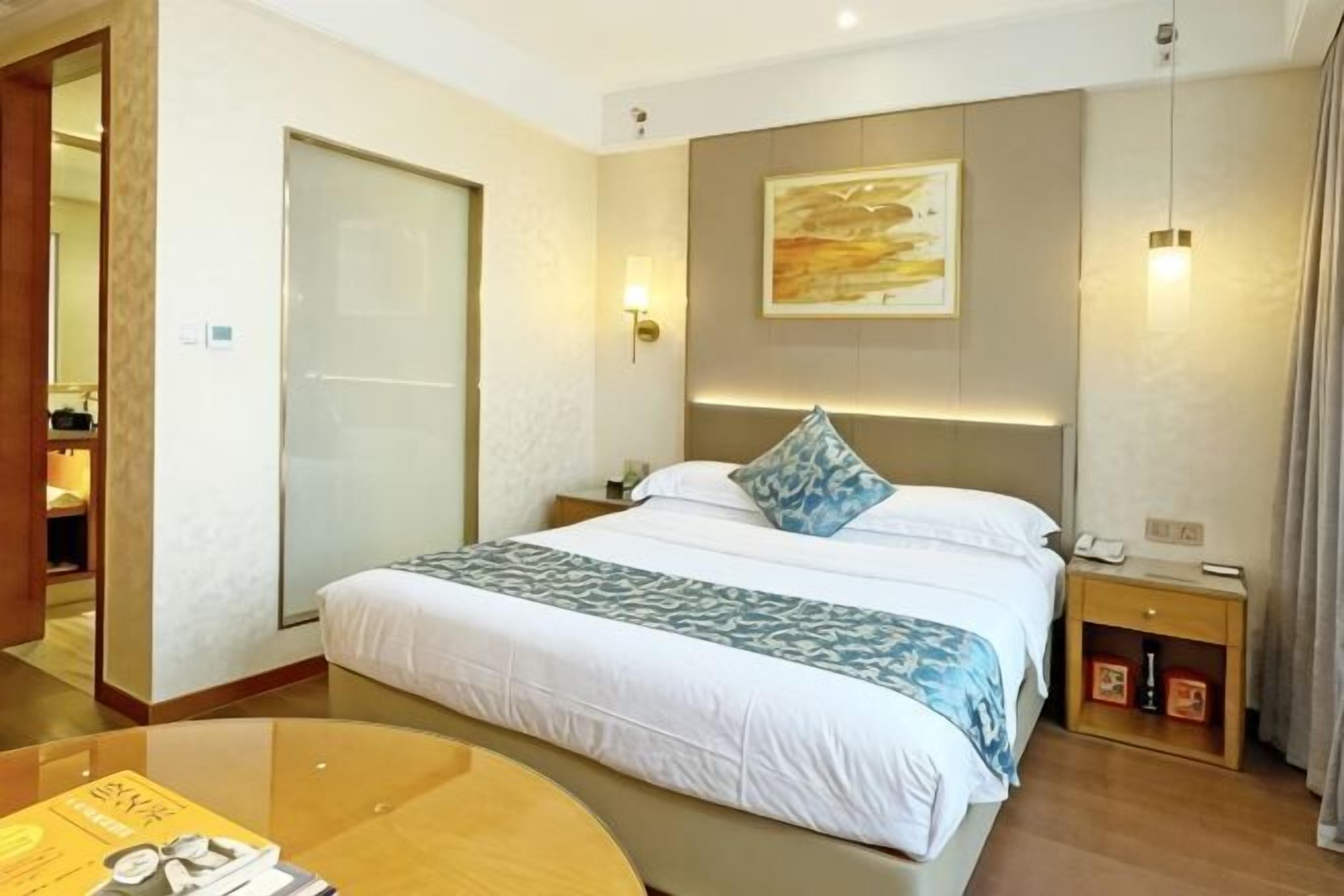 A hotel room with a bed, bedside table, lamp, and a painting on the wall.