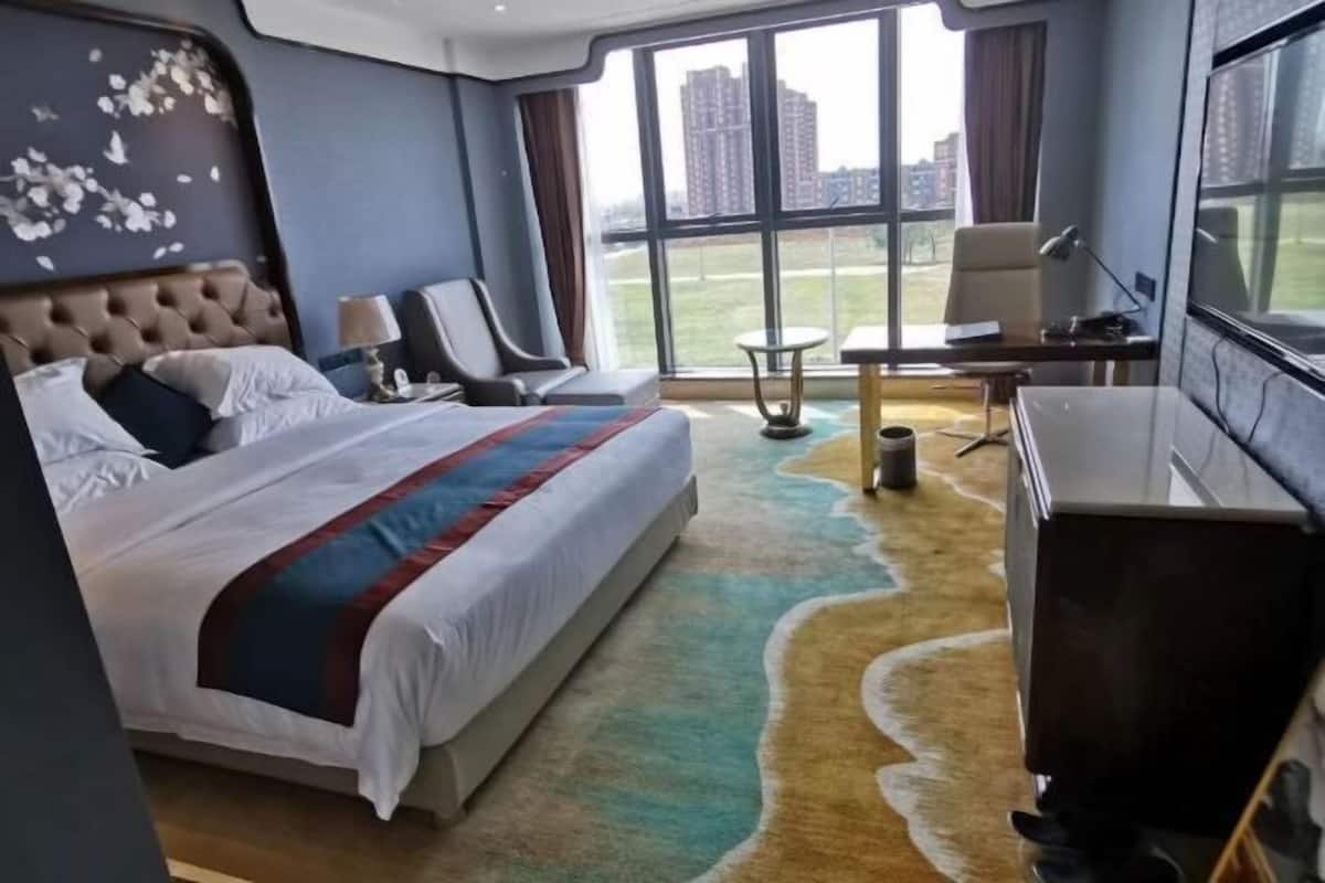 A hotel room with a large bed, a desk, a chair, and a view of the city through large windows.