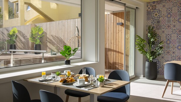 A modern dining area with a table set for breakfast, large windows, and potted plants. - Casual Raizes Porto Hotel by Casual Hoteles (Porto)