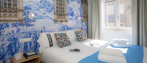 A hotel room with a large bed, a nightstand, a window with curtains, and a wall with blue and white decorative tiles.