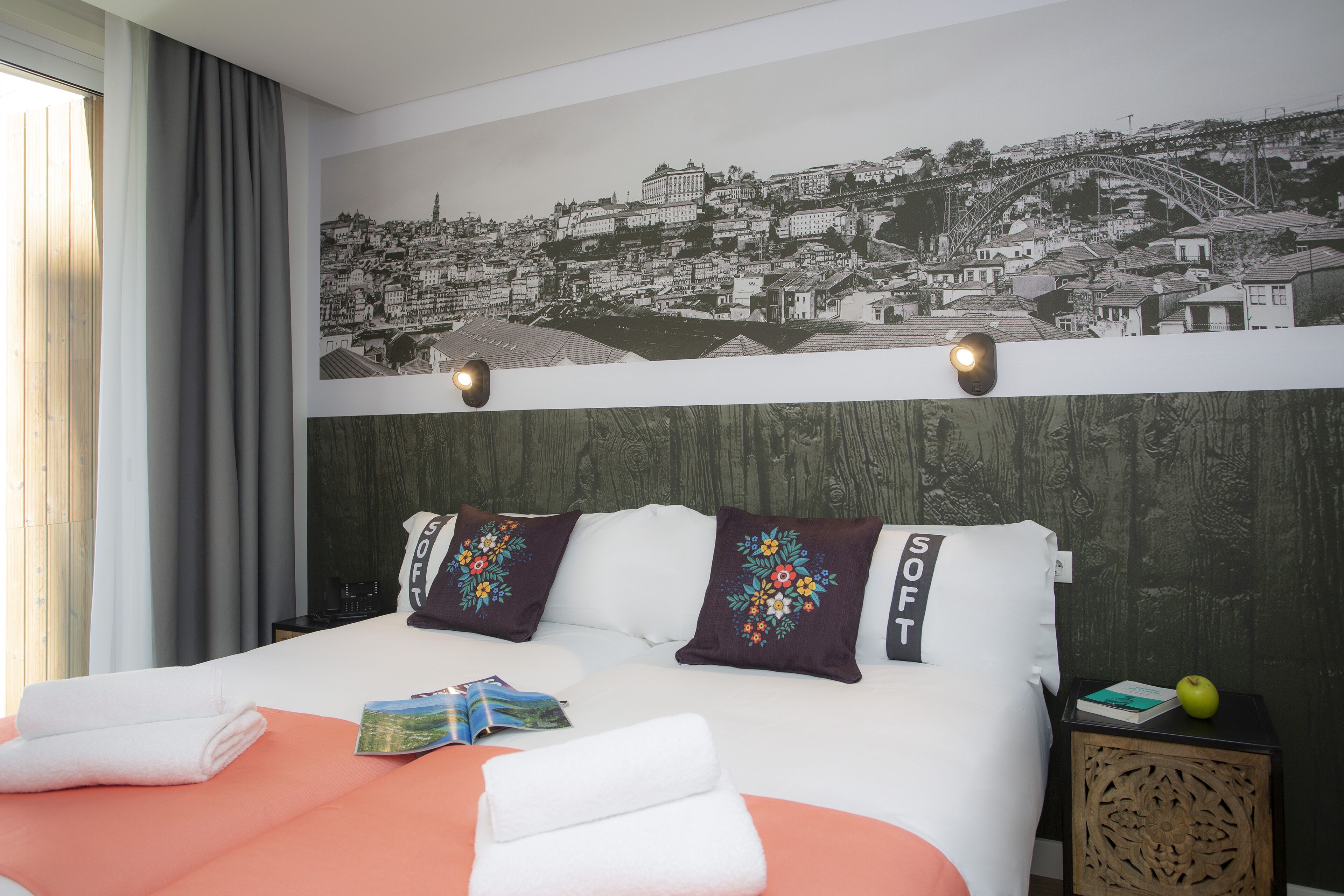 A hotel room with a bed, a nightstand, a wall-mounted lamp, and a cityscape mural.