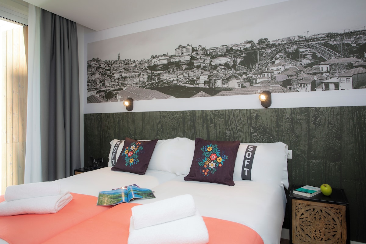 A hotel room with a bed, a nightstand, a wall-mounted lamp, and a cityscape mural.
