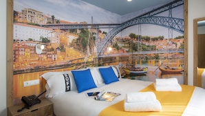 A hotel room with a mural of a cityscape and a bridge, a bed with white bedding and blue pillows, a bedside table with a phone, and a magazine. - Casual Raizes Porto Hotel by Casual Hoteles (Porto)