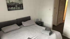 A neatly made bed with a grey headboard, two pillows, and rolled towels. A small bedside table with a lamp is visible. - Lovely 2-bed Apartment in Solihull (Solihull)