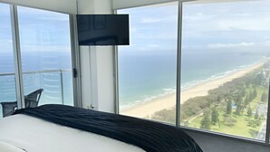 A bedroom with a large bed, a TV mounted on the wall, and a view of the beach and ocean through floor-to-ceiling windows.