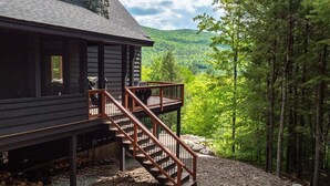 A cabin with a wooden exterior, a stone chimney, a wooden staircase, and a view of a forested mountain. - Luxury Chalet | Hot Tub | Minutes to Sunday River (Newry)