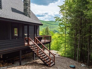 A cabin with a wooden exterior, a stone chimney, a wooden staircase, and a view of a forested mountain. - Luxury Chalet | Hot Tub | Minutes to Sunday River (Newry)