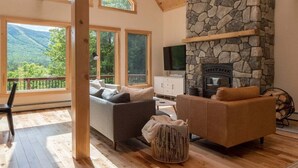 A cozy living room with a stone fireplace, wooden beams, and large windows overlooking a mountainous landscape. - Luxury Chalet | Hot Tub | Minutes to Sunday River (Newry)