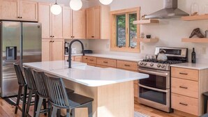 A modern kitchen with wooden cabinets, a stainless steel refrigerator, a gas stove, and a large island with bar stools. - Luxury Chalet | Hot Tub | Minutes to Sunday River (Newry)