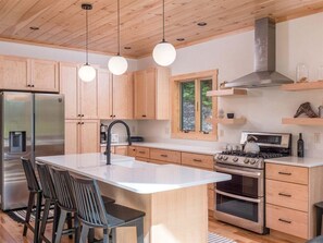 A modern kitchen with wooden cabinets, a stainless steel refrigerator, a gas stove, and a large island with bar stools. - Luxury Chalet | Hot Tub | Minutes to Sunday River (Newry)