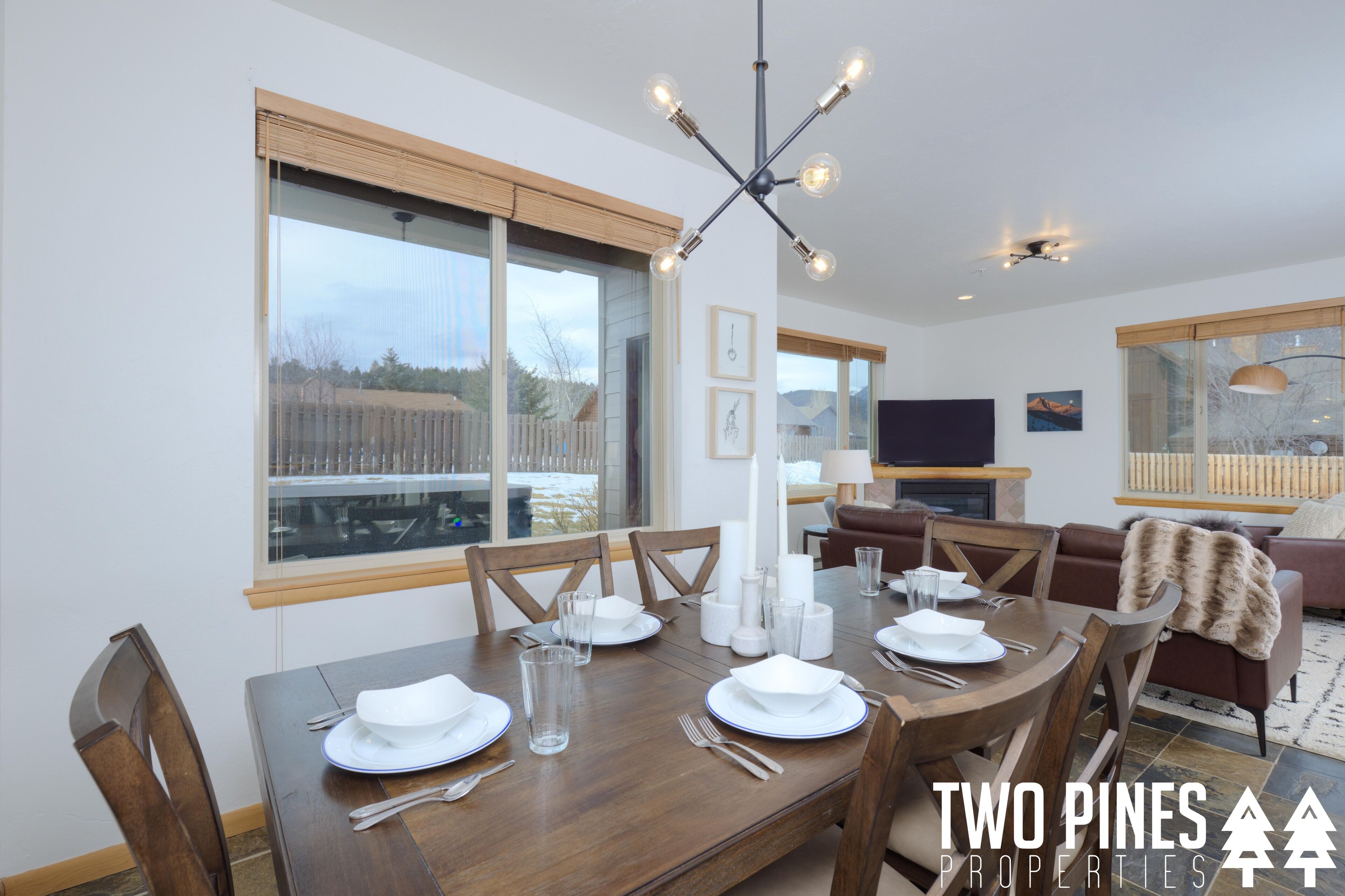 A dining area with a wooden table set for four, a chandelier, and large windows with a view of a wooden deck.