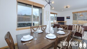 Een eethoek met een houten tafel gedekt voor vier personen, een kroonluchter en grote ramen met uitzicht op een houten terras.