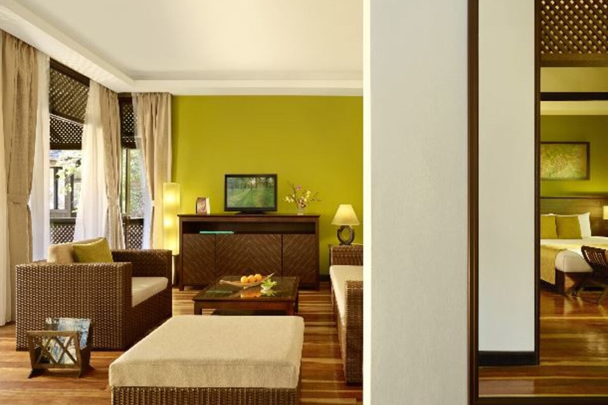 A hotel room with a green wall, wicker furniture, a flat-screen TV, and a dining table with fruit.