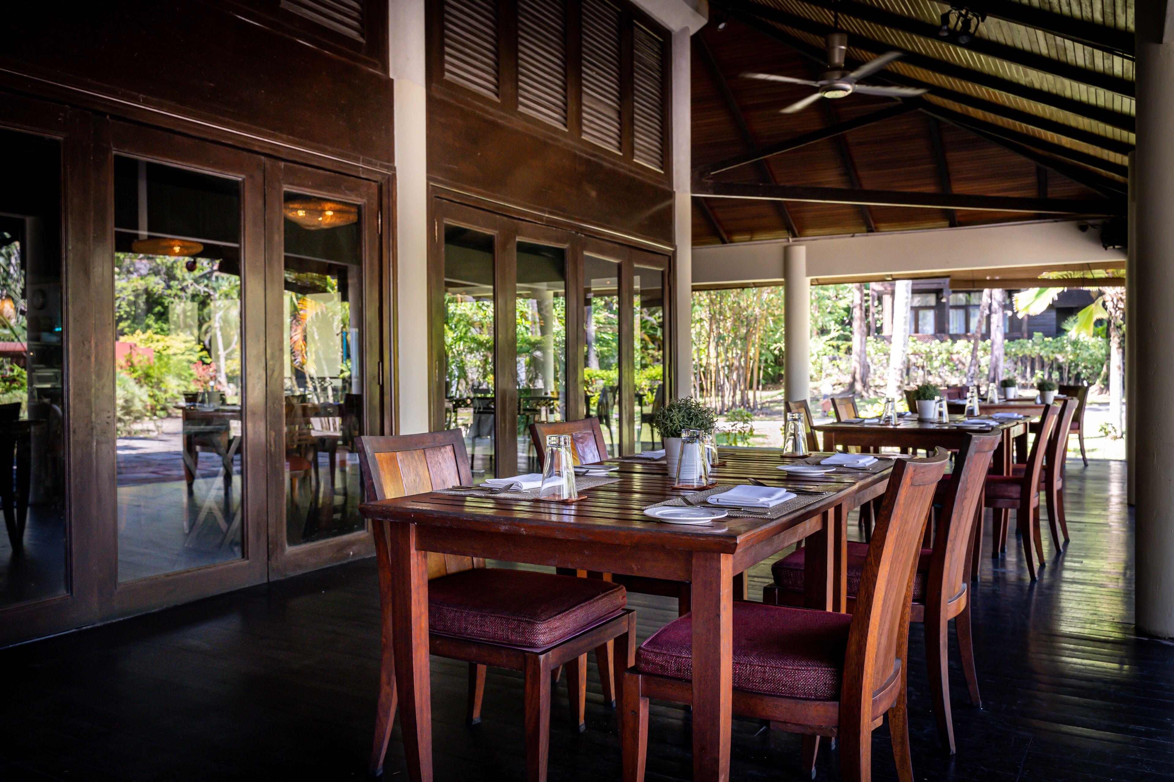 A restaurant with wooden tables and chairs, large windows, and a ceiling fan.