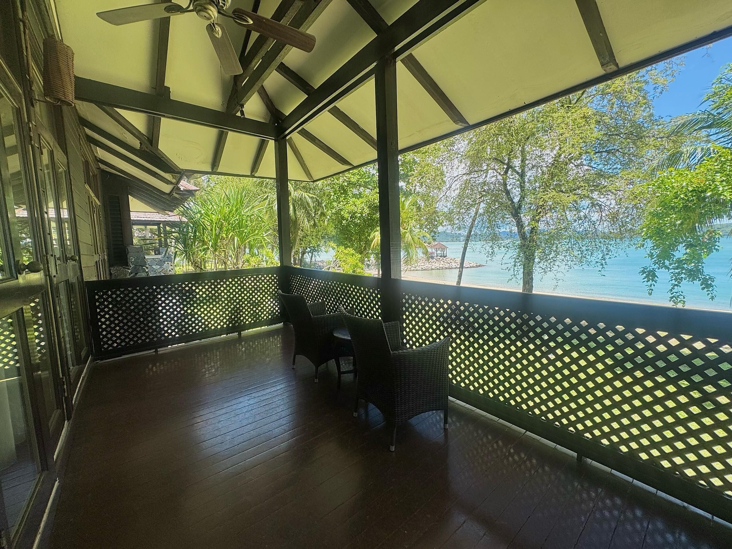 A covered outdoor area with a wooden floor, a ceiling fan, and a view of a beach and trees.